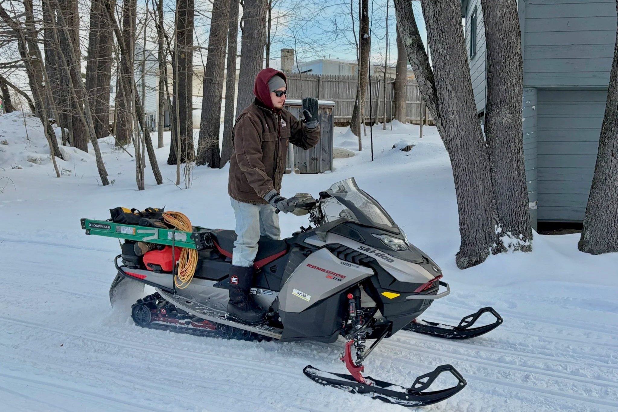 Man standing on a snowmobile in a snowy wooded area, wearing a brown jacket, gray pants, gloves, and sunglasses, raising his hand in a wave.
