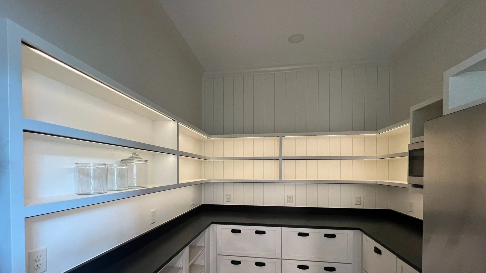 Empty white kitchen with black countertops, open white cabinets, and glass jars on the shelves.