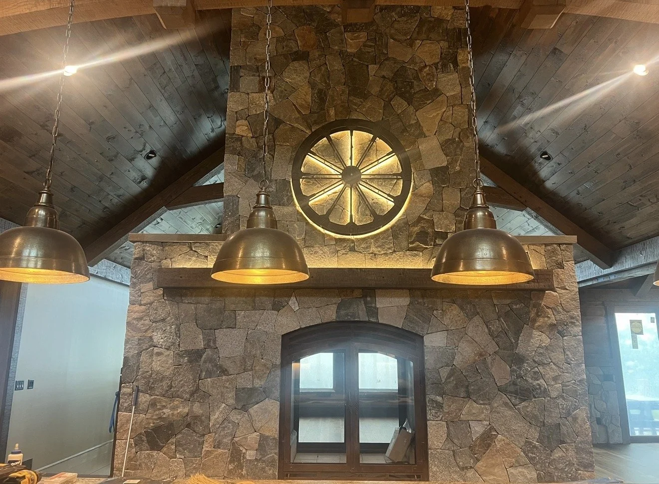 Interior of a house with a stone fireplace, a large round light fixture resembling a wheel, and three hanging metallic pendant lights.