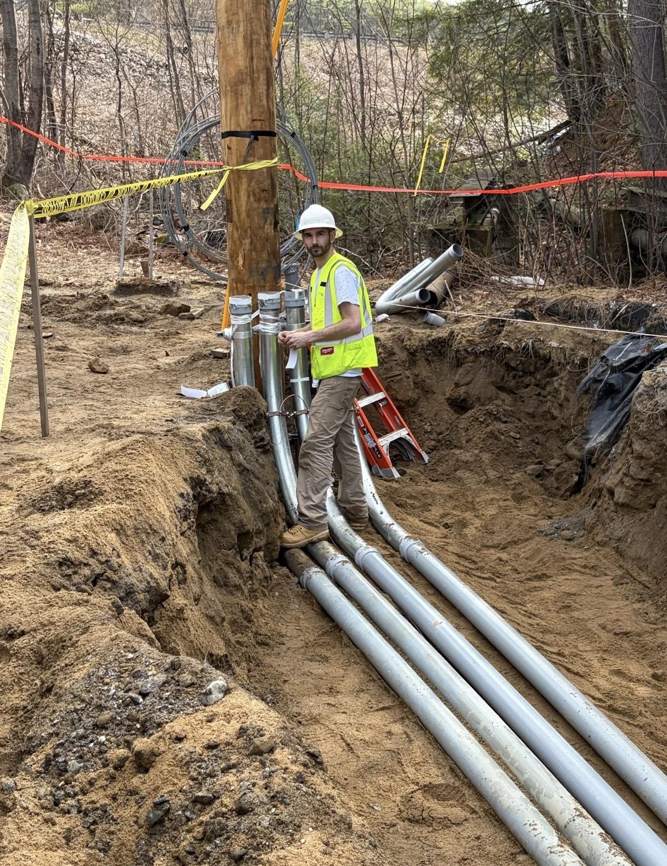 A construction worker wearing a white hard hat and yellow safety vest is standing next to large pipes in a dug trench, with construction materials and warning tape around the site.