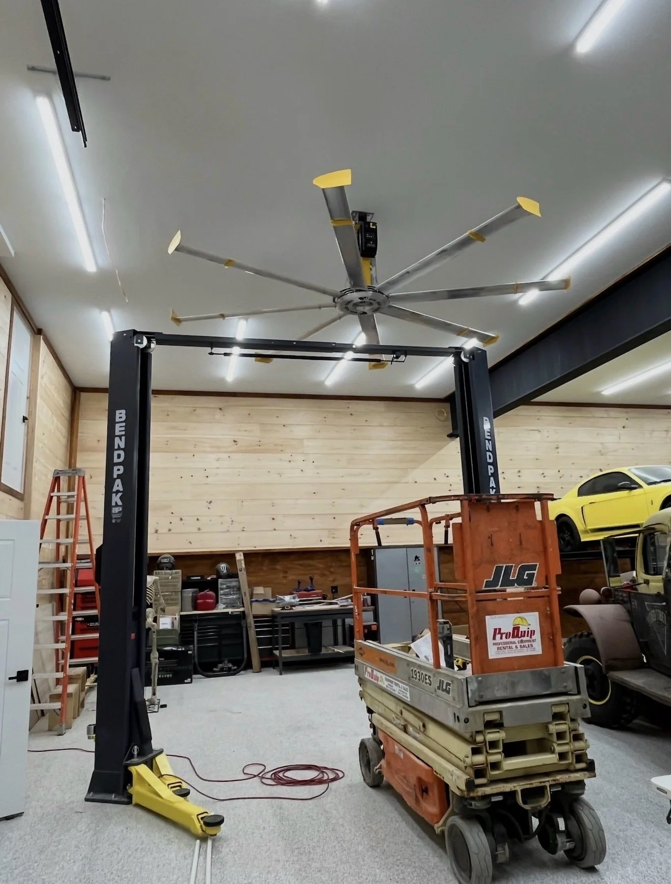 Garage or workshop with equipment including a black lift, orange lift, and yellow tool, and a large ceiling fan with yellow blades.