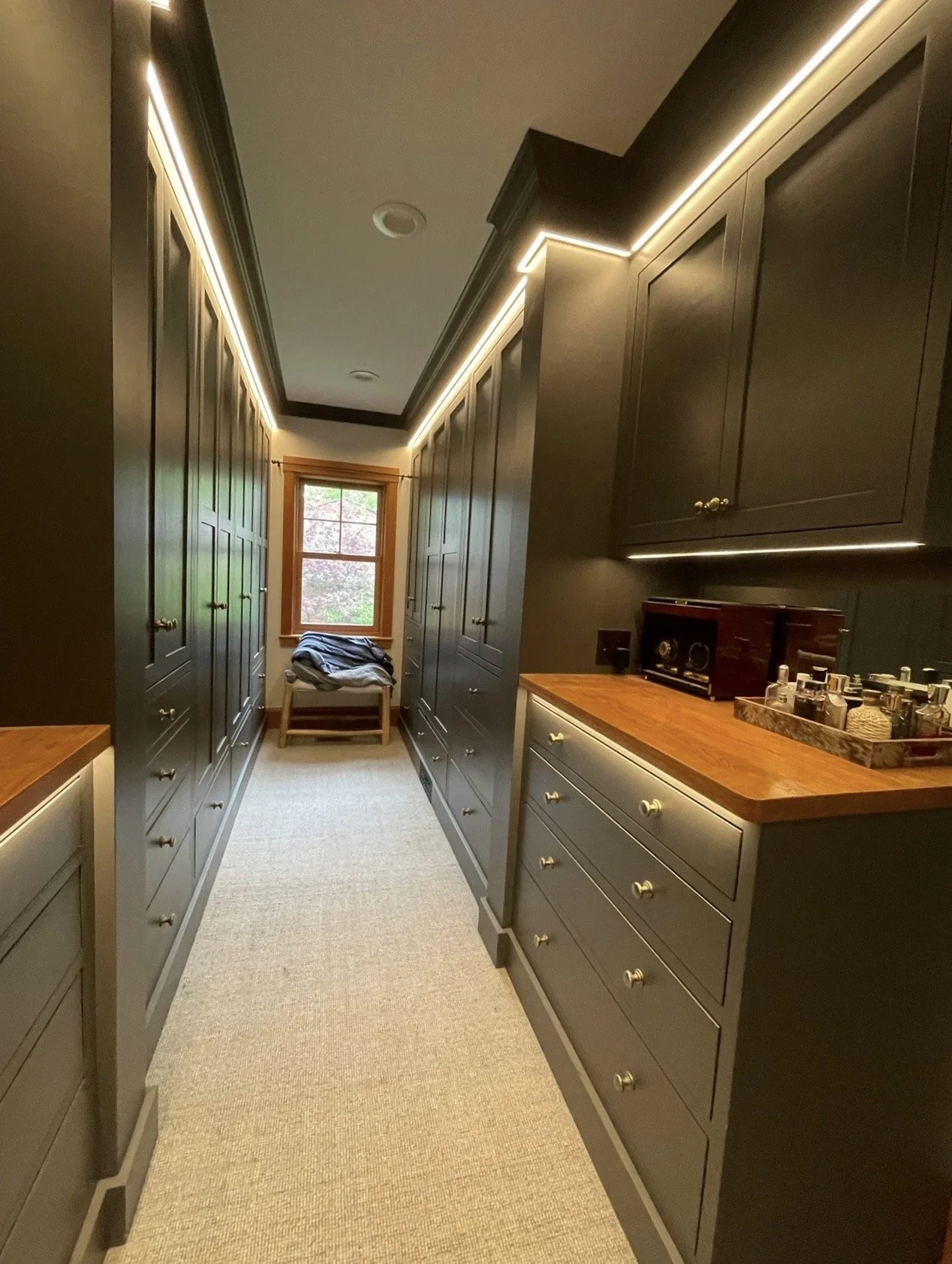 A narrow walk-in closet with dark gray built-in cabinets and drawers on both sides, a beige carpeted floor, and a window at the end with a small bench beside it holding clothes. The ceiling has recessed lighting with LED strip accents.