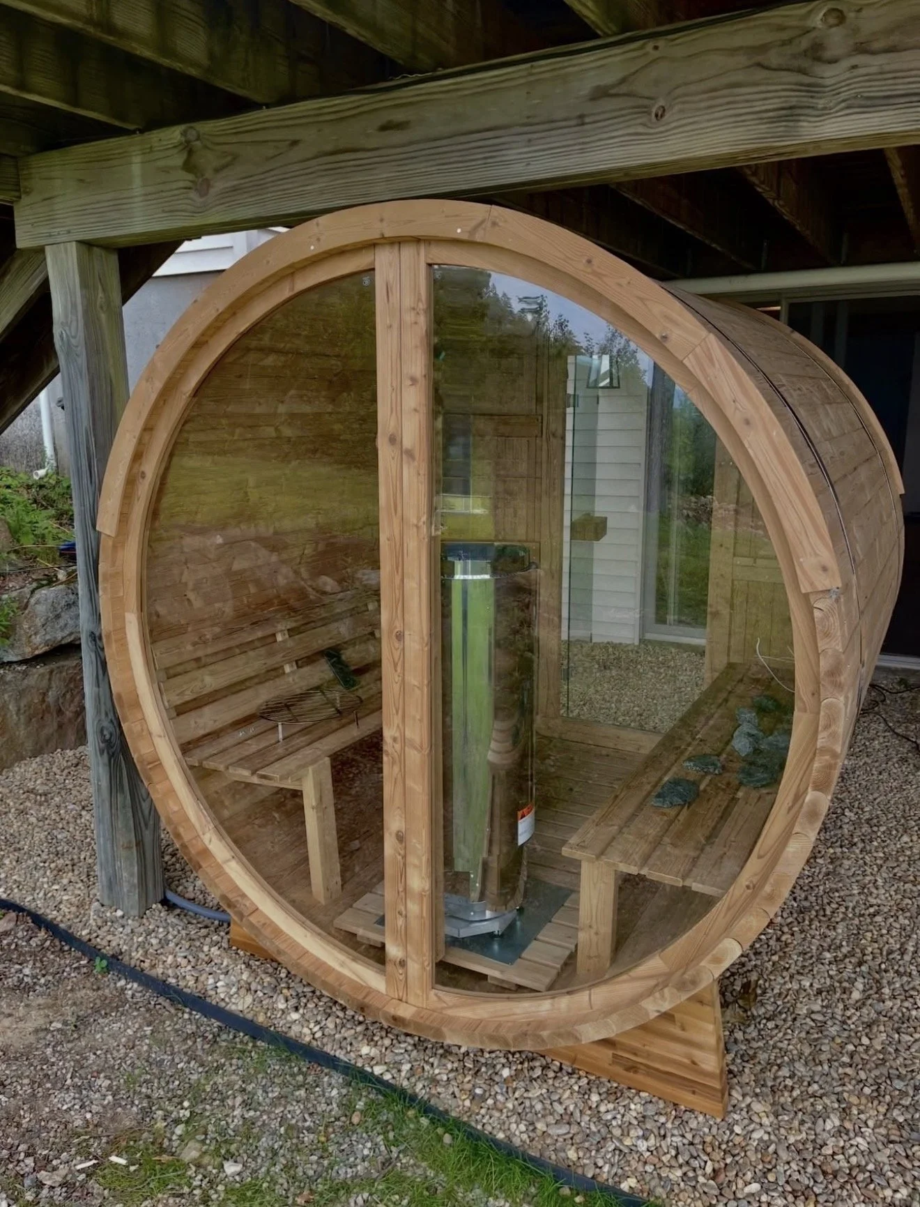 A small, round wooden sauna with glass walls and doors, located outdoors on gravel, built under a wooden deck.