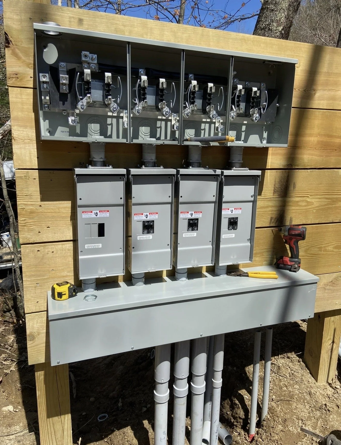 Electrical meter box with circuit breakers and electrical wiring mounted on a wooden panel outdoors.
