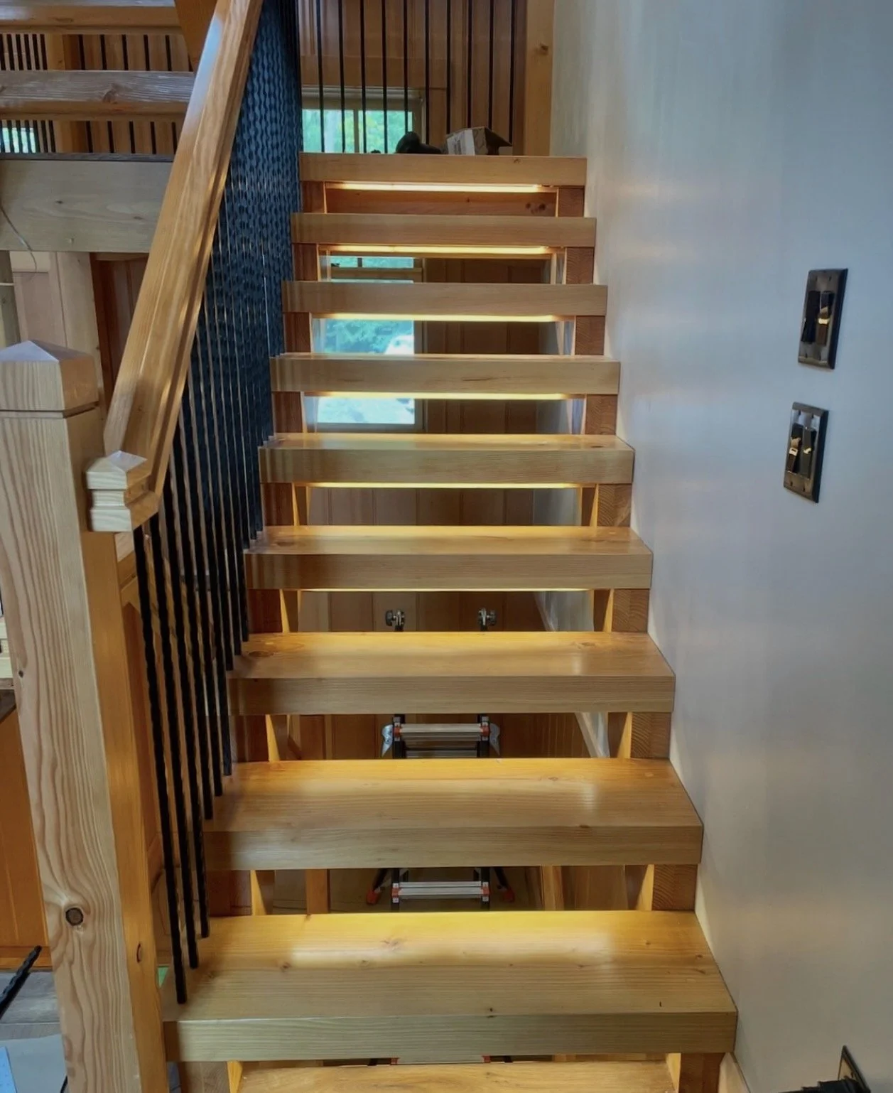 Unfinished wooden staircase with treads, railing and a black spindled guard on the left side, against a cream-colored wall with two electrical outlets on the right, during construction.