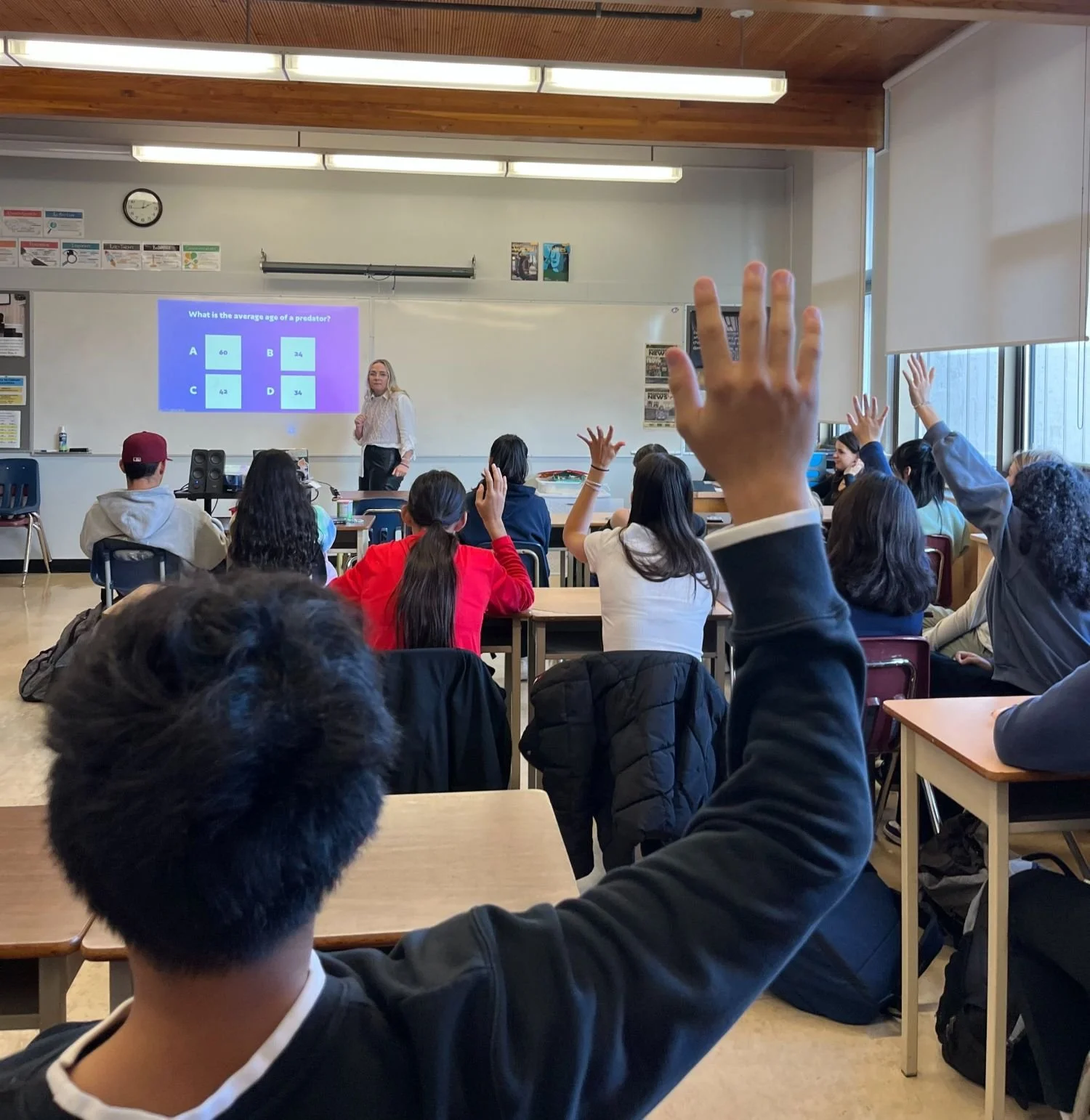A classroom with students raising their hands, facing a teacher who is standing at the front near a whiteboard with a lesson projected on it. The classroom has large windows on the right side and posters on the walls.