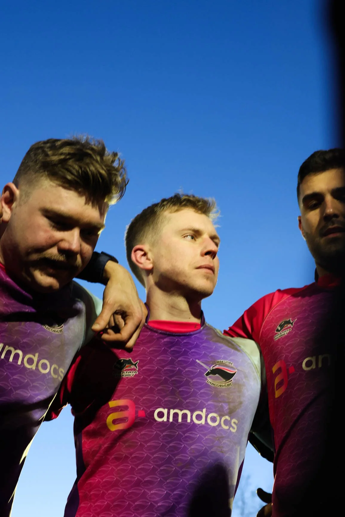 Three rugby players in maroon and purple jerseys with sponsorship logos, standing close together with their arms around each other, against a clear blue sky.