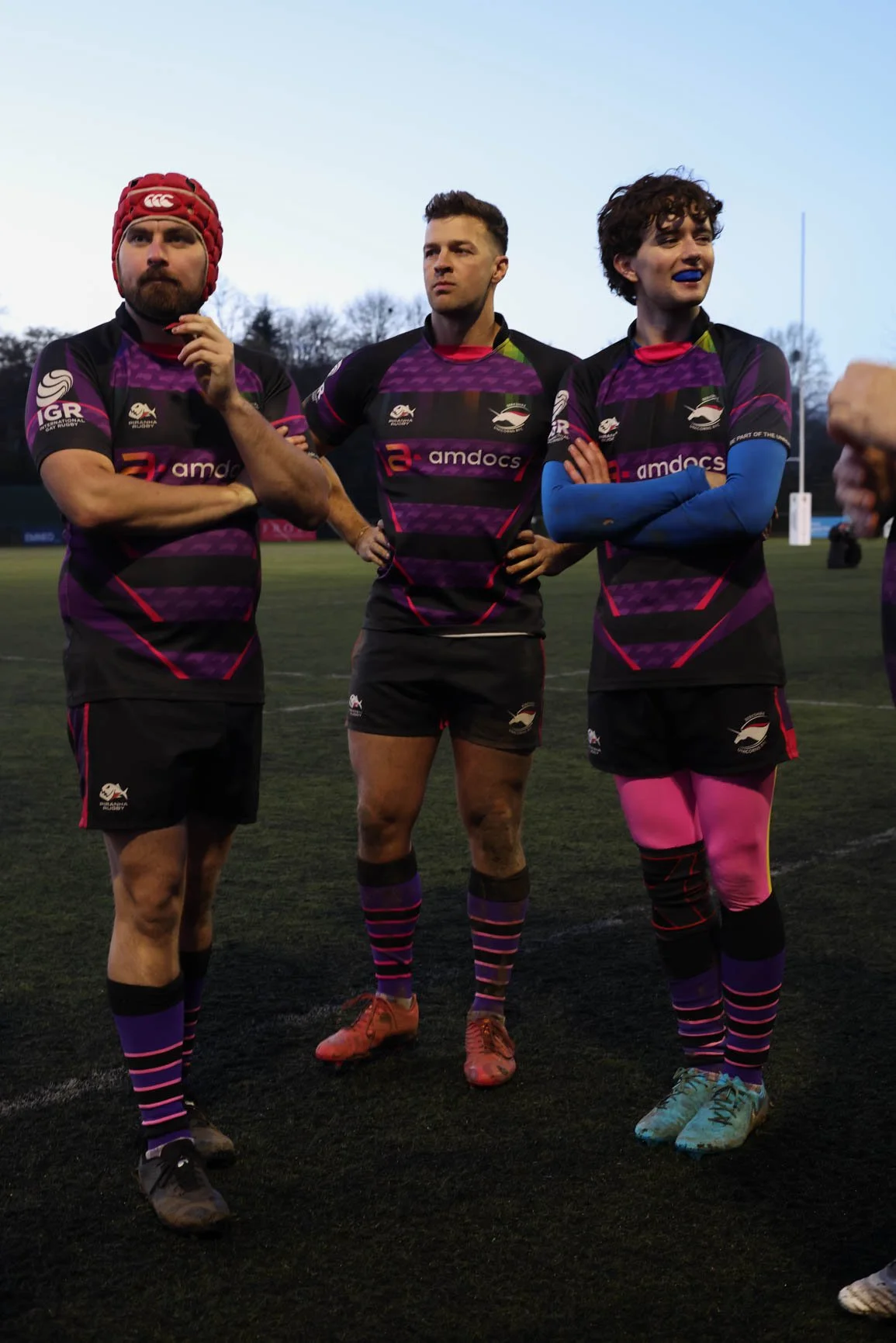 Three rugby players standing on a field during dusk, wearing black and purple uniforms with pink and blue accents, with trees in the background.
