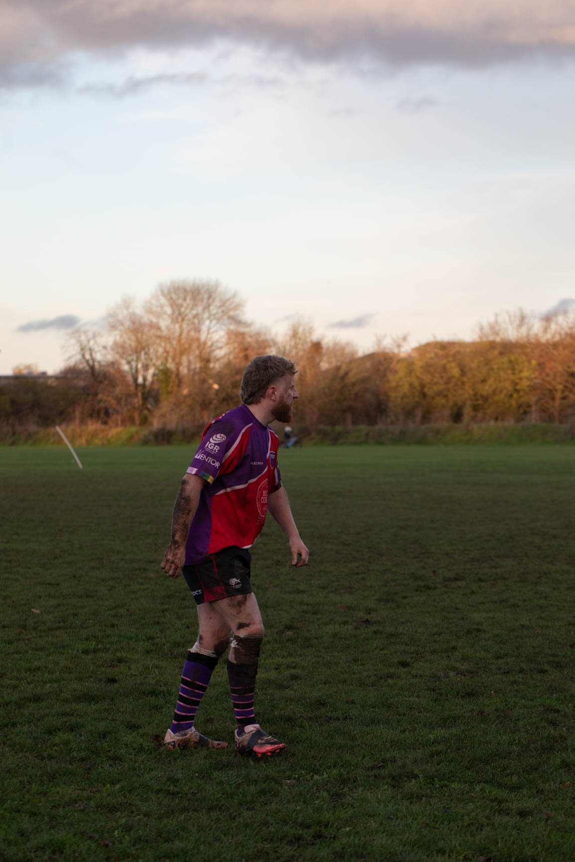 A rugby player walking on a grassy field with mud on his legs, wearing a purple and red jersey, black shorts, and purple striped socks, with trees and a cloudy sky in the background.