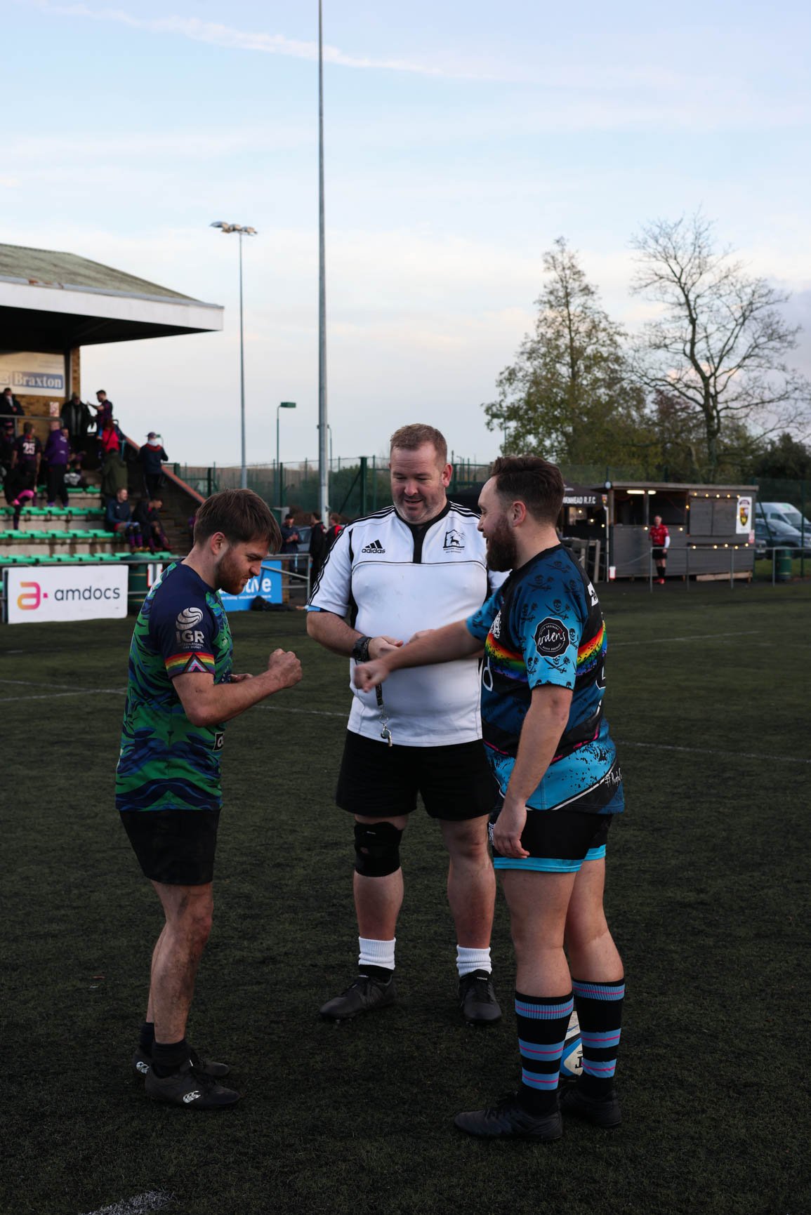 Two rugby players, one in a blue and teal jersey and the other in a rainbow-colored jersey, are interacting with an official in a white rugby uniform on a rugby field. The official appears to be making a decision while the players are discussing, wit