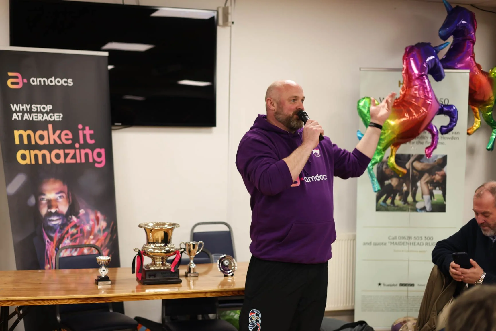A man wearing a purple hoodie, holding a microphone, and speaking at an indoor event with colorful balloon horses in the background. There are trophies on a table nearby, and a banner promoting 'amdocs'.