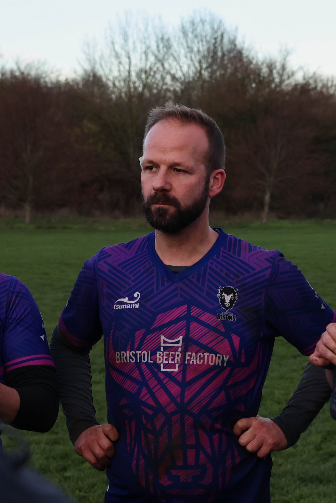 Man with a beard and short brown hair in a purple sports jersey standing outdoors with trees in the background.