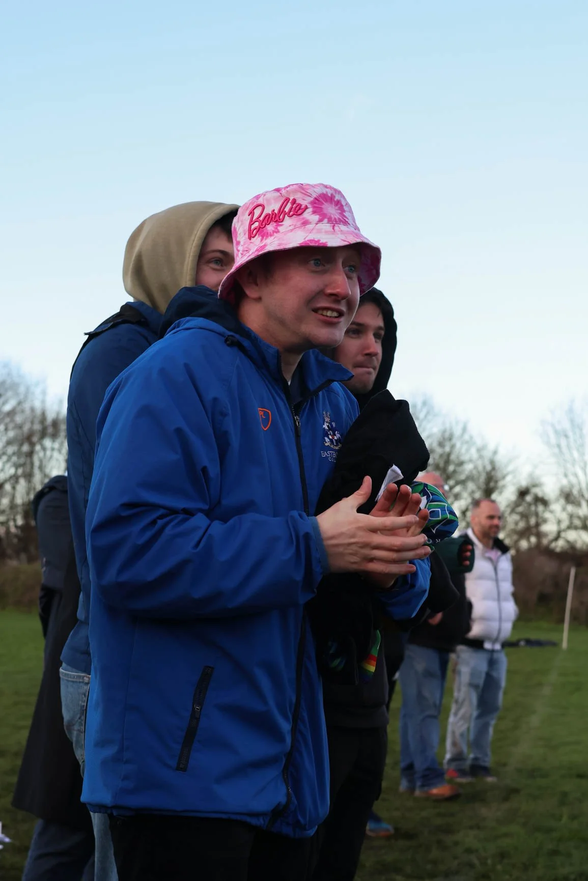 A group of people outdoors during daytime, wearing jackets and hats, with the main focus on a young man wearing a pink 'Barbie' bucket hat, smiling, holding a black item, standing on a grassy field with trees in the background.