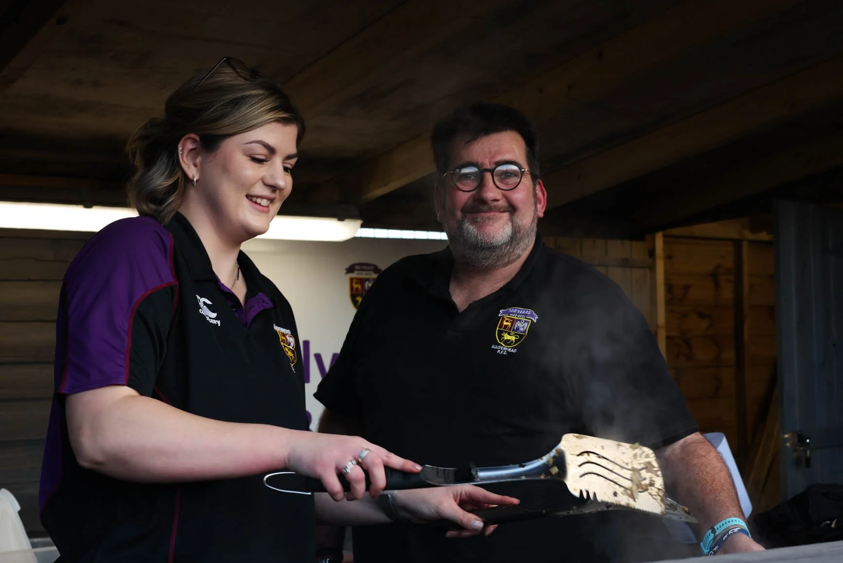 A woman and a man smiling as they hold a BBQ spatula and cook food outdoors, wearing black shirts with a badge logo, in a wooden shed or pavilion.