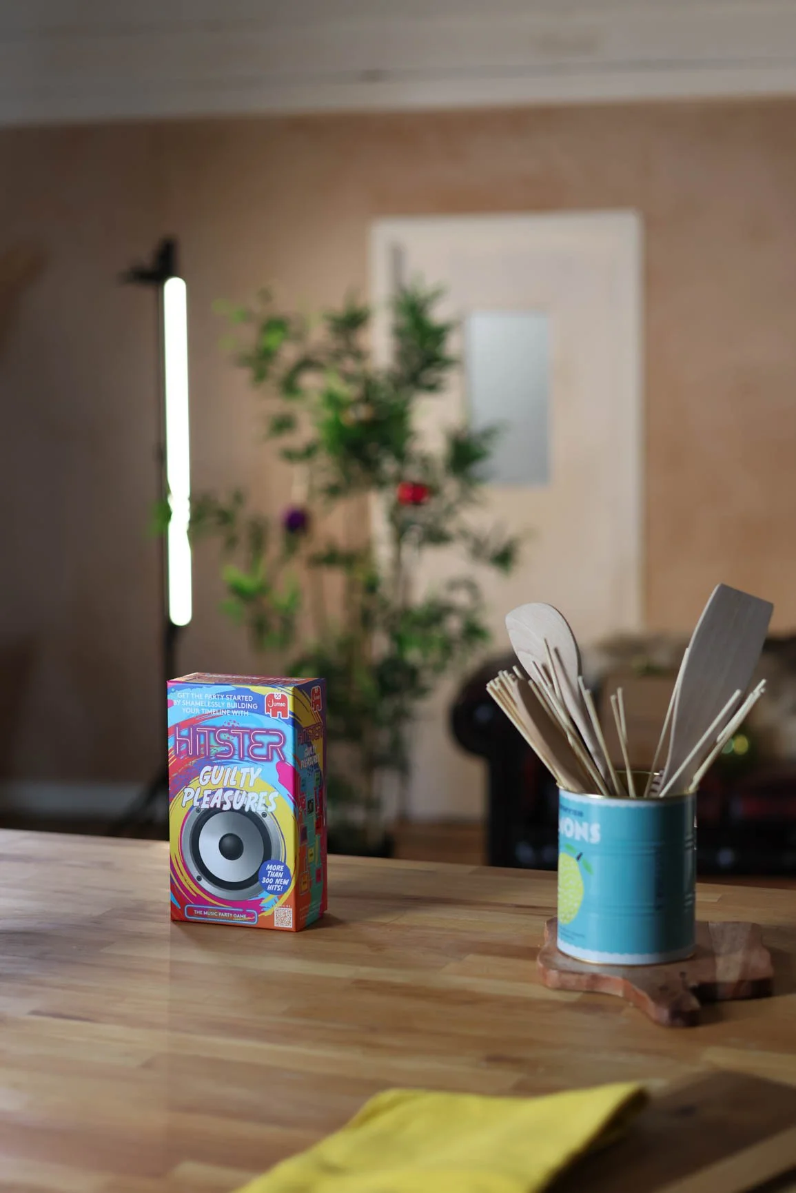 Party game box labeled "Hyster Guilty Pleasures" on a wooden table, with a container holding utensils or paper products, and a blurred background with a tall plant and wall-mounted lighting.