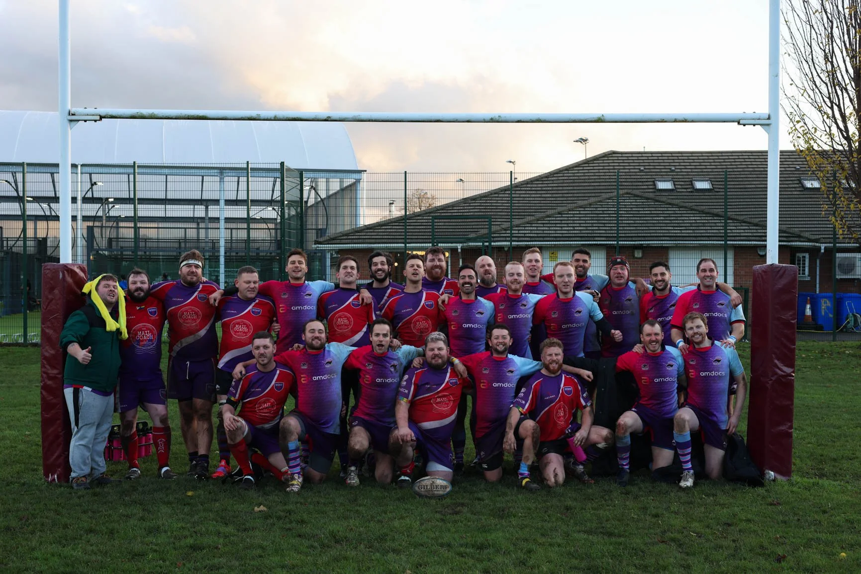 A group of rugby players and team staff posing for a photo on a rugby field with goal posts, a fence, and buildings in the background.