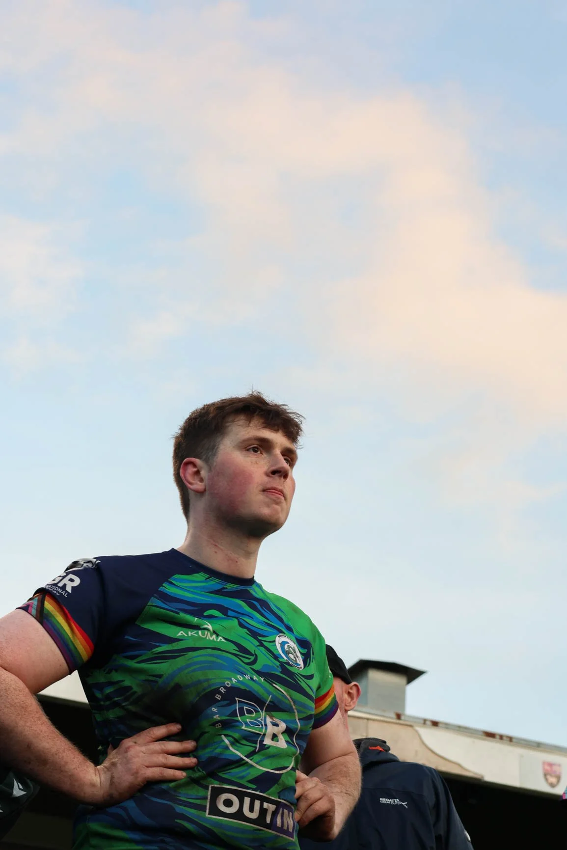 A young male athlete stands outdoors with a blue sky and clouds above. He has short brown hair and is wearing a colorful sports jersey with rainbow accents.