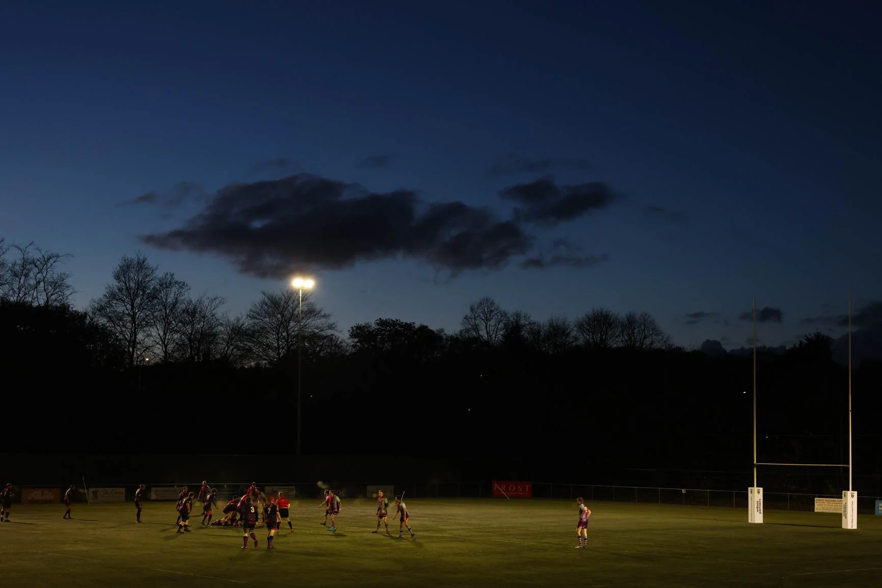 Rugby game played at dusk on a field with players in dark and light jerseys. Goal posts on the field, and stadium lights illuminating from above. Dark sky with clouds and trees in the background.