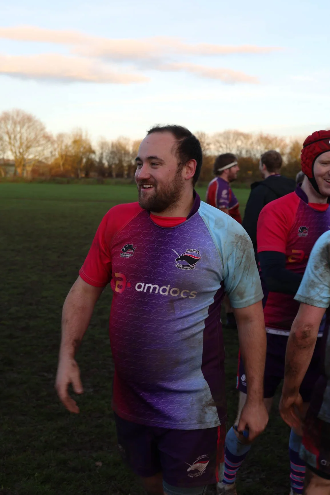 A group of rugby players, wearing maroon and purple jerseys with the logo of Berkshire Unicorns RFC and amdocs, standing on a grassy field with trees and a cloudy sky in the background. One player in the foreground is smiling, with mud on his arm.