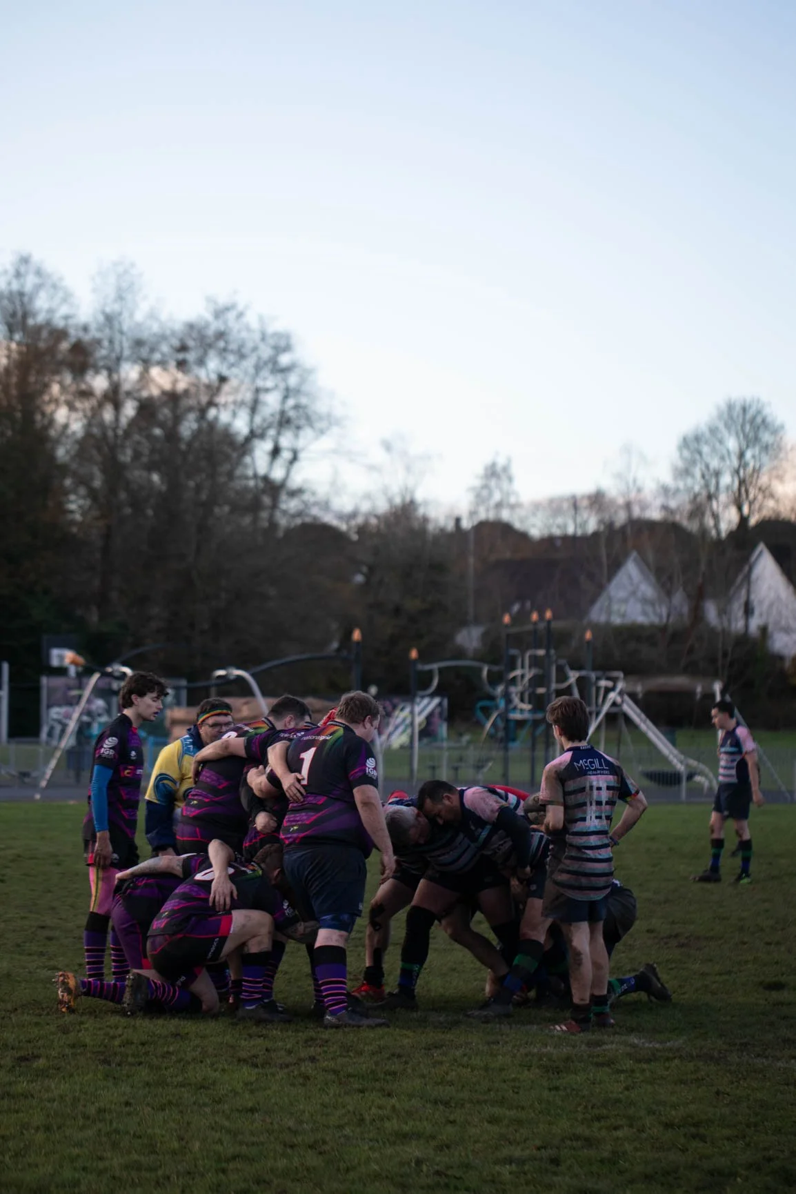 A rugby match taking place on a grassy field during late afternoon or early evening, with players engaged in a scrum and others standing around. In the background, there are trees, houses, and a playground.