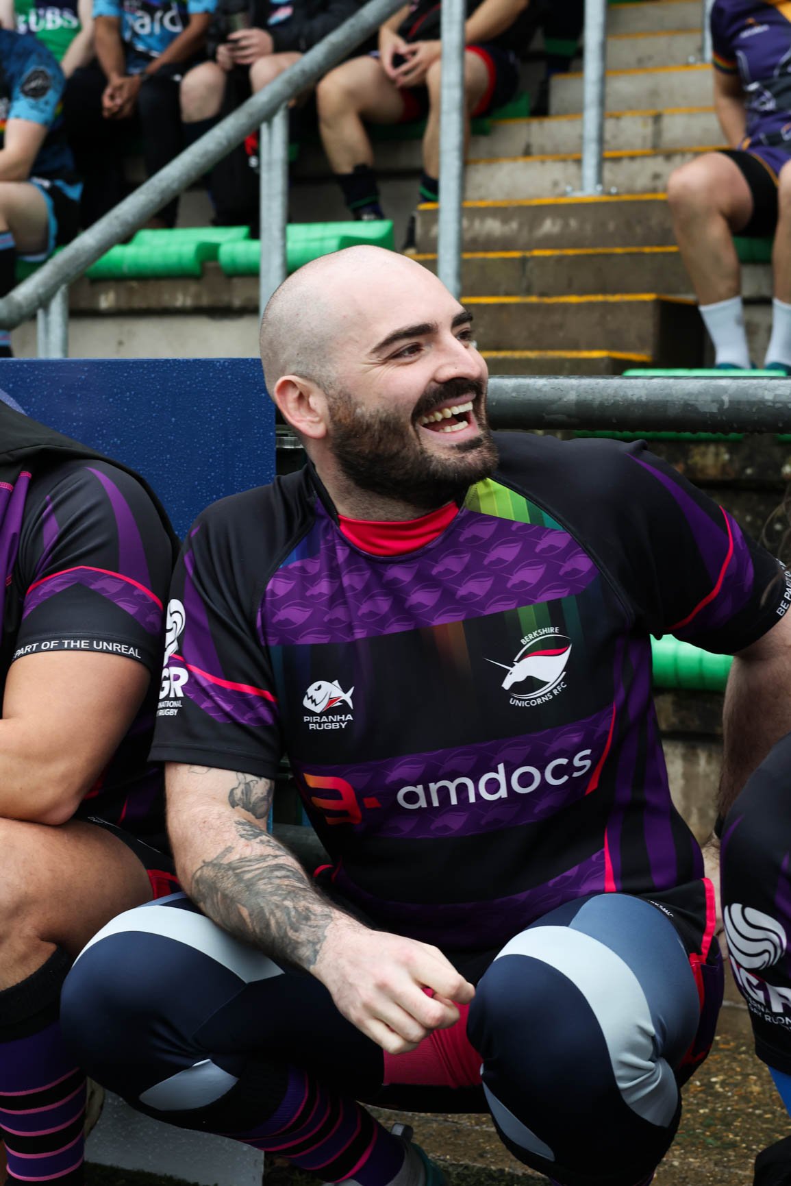 A smiling rugby player with a beard and tattoos, wearing a purple and black jersey with logos, sitting on a bench with other players and spectators in the background.