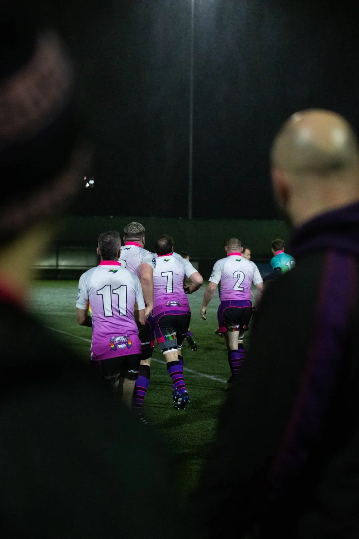 Rugby players in purple and white uniforms running onto the field at night, seen through a gap in the crowd.