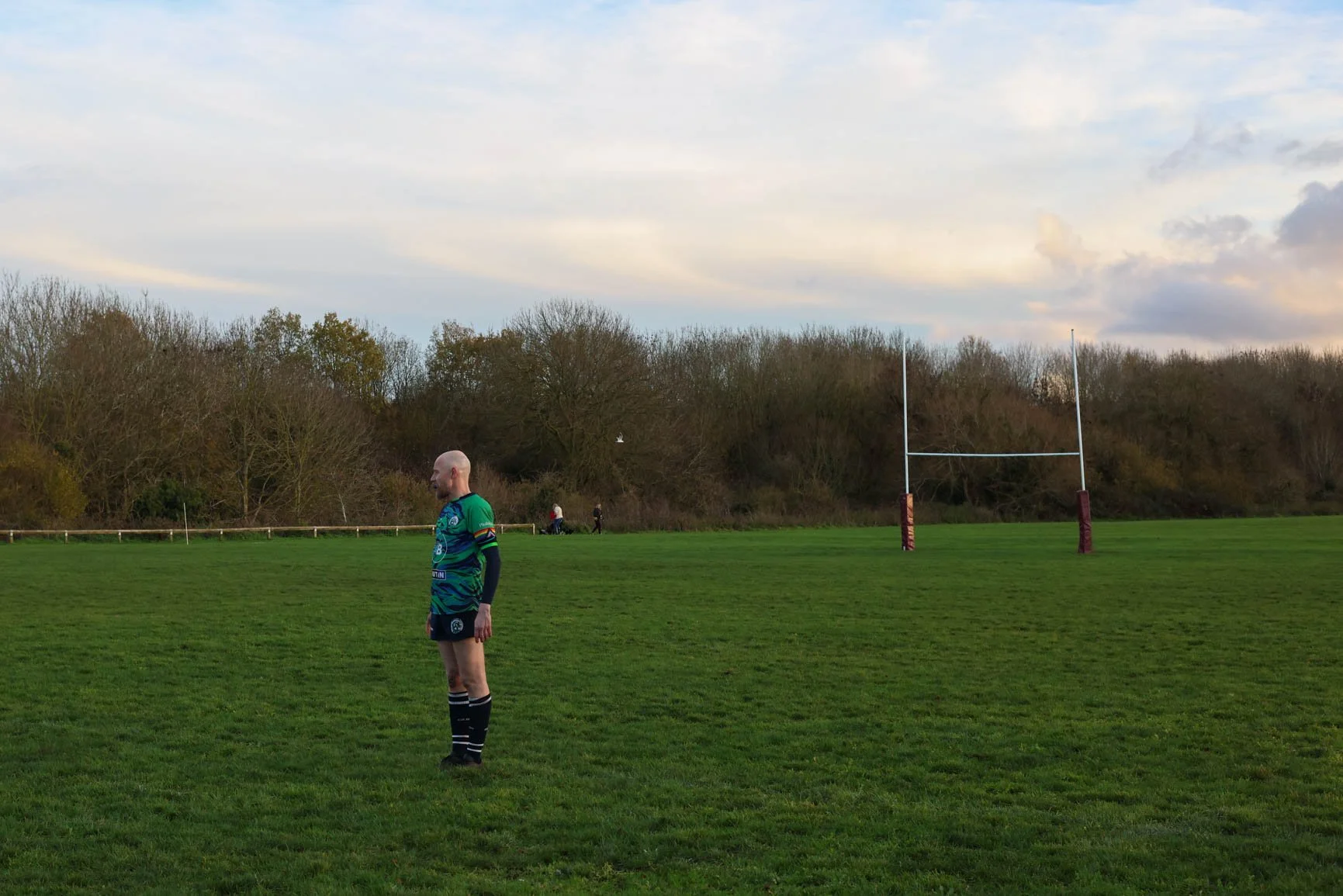 A rugby player standing on a grassy field near a goal post, with trees and a cloudy sky in the background.