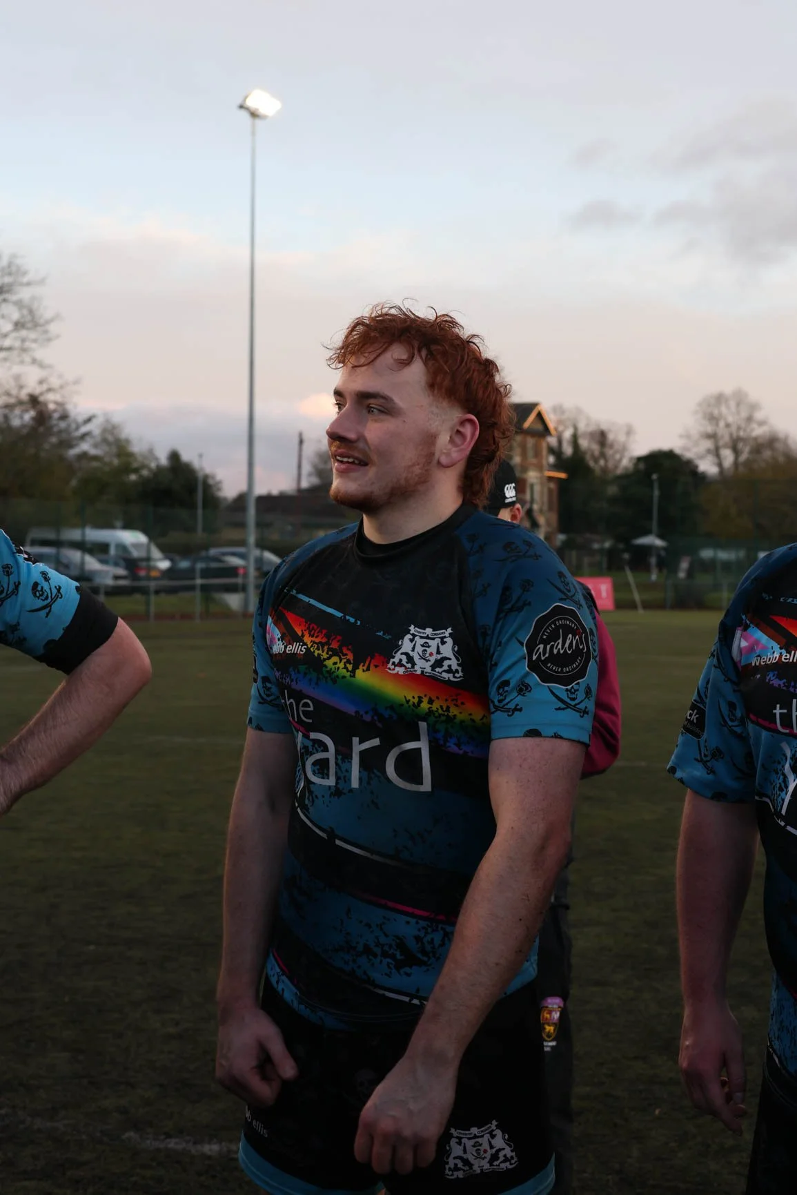 A young man with curly red hair and light skin wearing a black rugby jersey with rainbow-colored stripes and black shorts, standing on a grassy field during sunset with a few other players partially visible nearby.