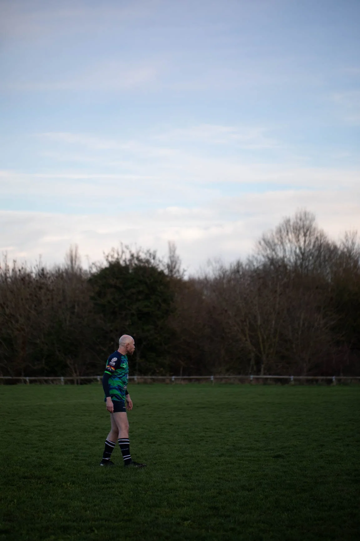 A man in a sports uniform standing on a grassy field with trees and a cloudy sky in the background.