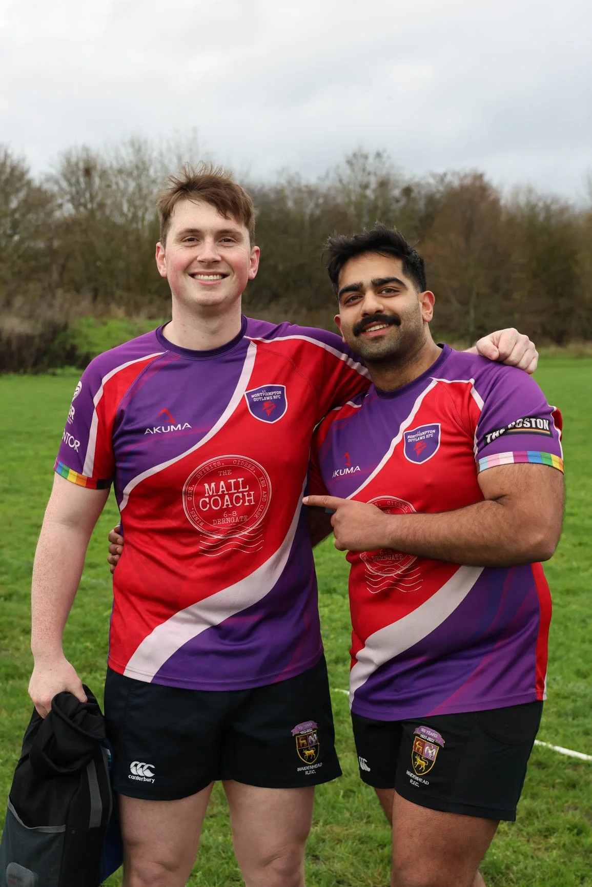 Two men standing outdoors on a grassy field, wearing matching purple and red rugby jerseys with team logos, smiling, with their arms around each other's shoulders.
