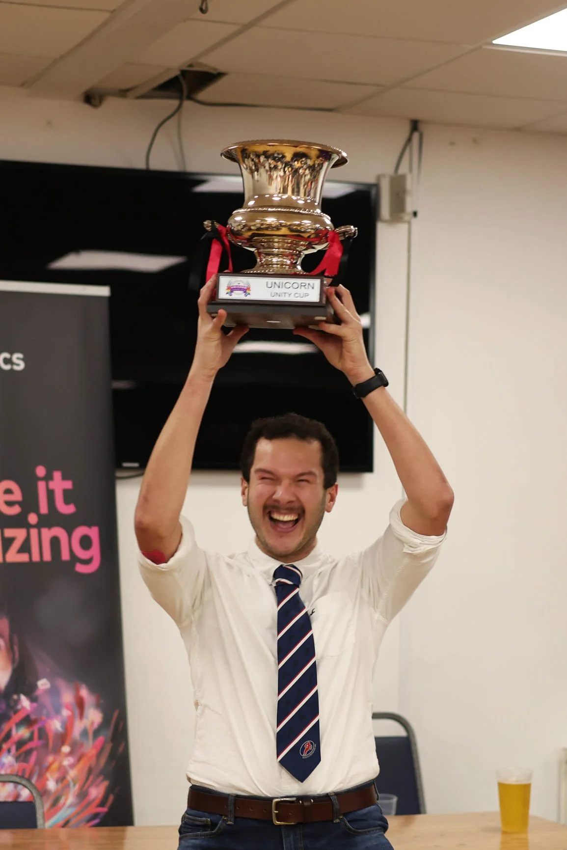 A man is smiling and holding a trophy above his head in a celebratory manner. The trophy is labeled 'Unicorn Unity Cup.' The man is wearing a white shirt, a striped tie, and a watch. There is a beer glass on the table in front of him and a poster wit