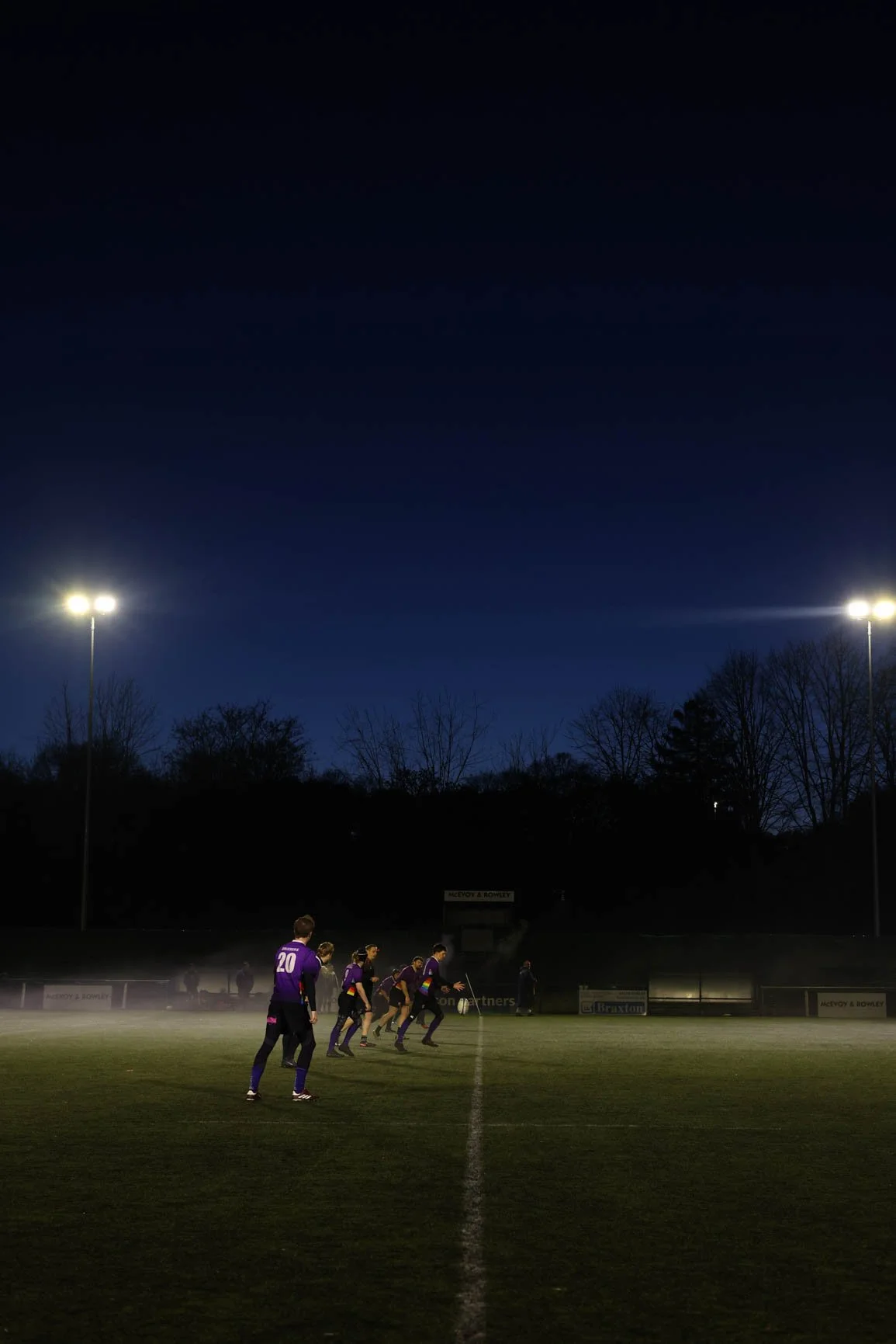 A soccer team dressed in purple jerseys warming up on a soccer field under stadium lights at night, with trees and a dark sky in the background.