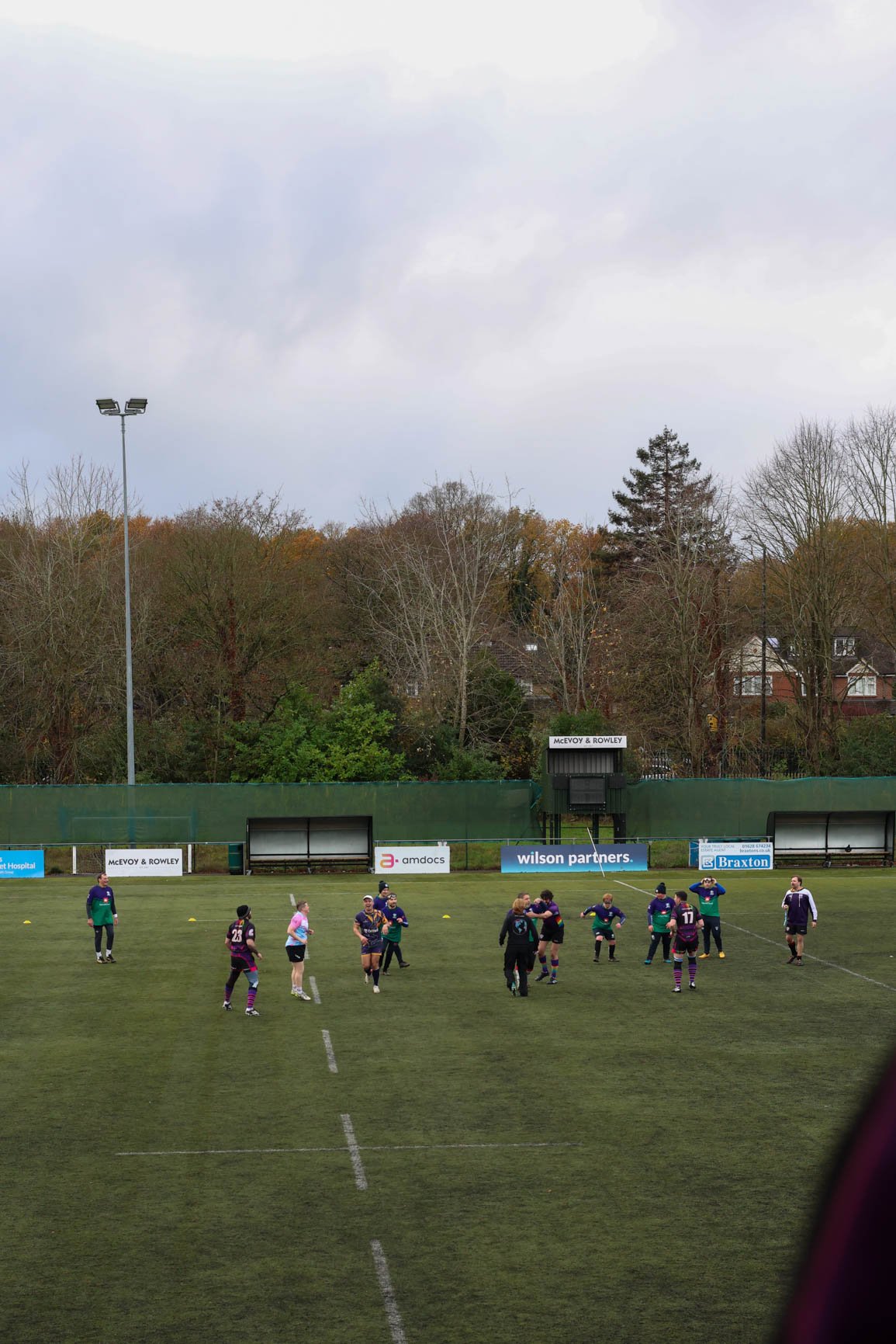A group of rugby players practicing on a field during daytime, with spectators watching from the sidelines, surrounded by trees and houses in the background.