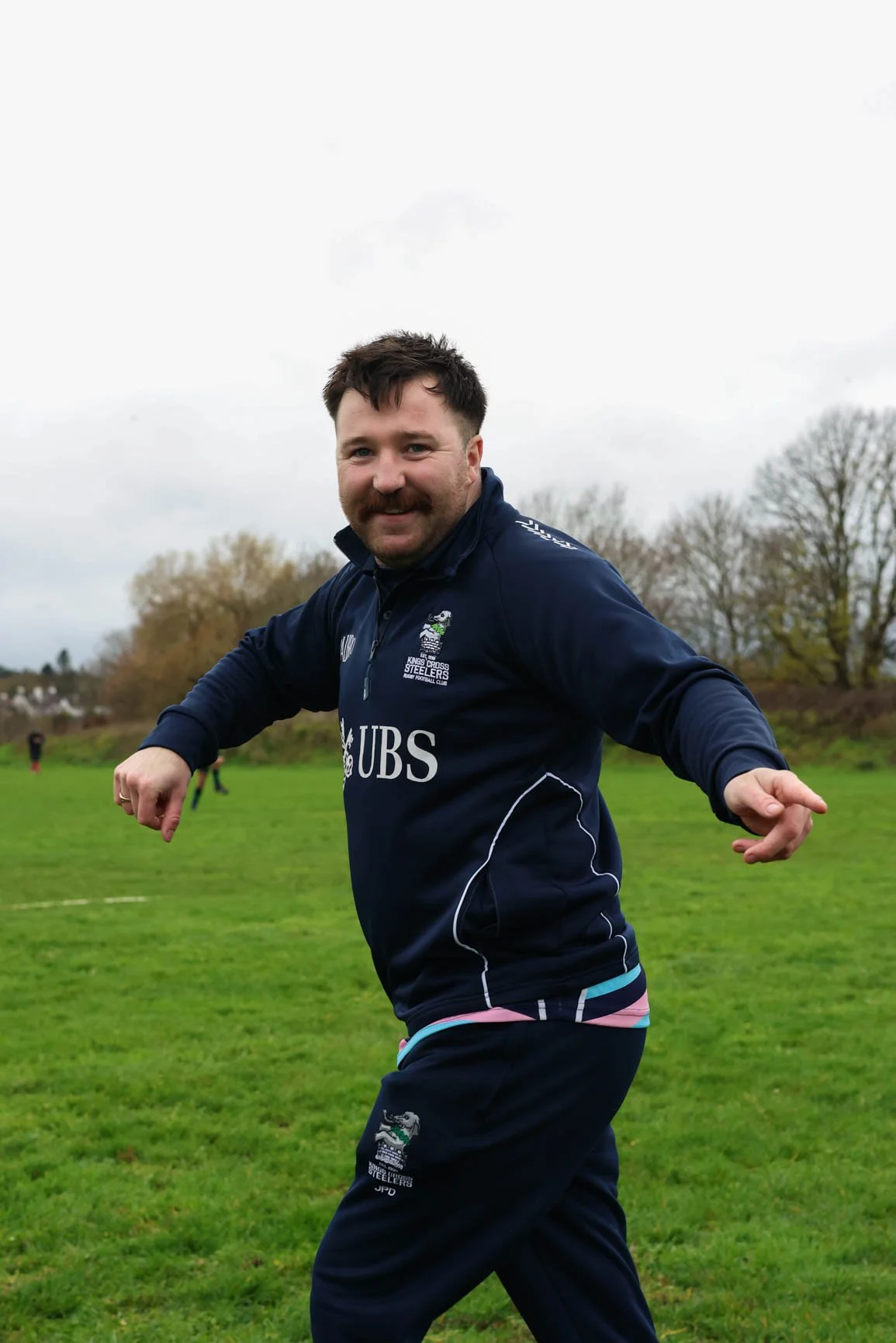 A man wearing a navy blue sports jacket with logos, smiling and running outdoors on a grassy field with trees and cloudy sky in the background.