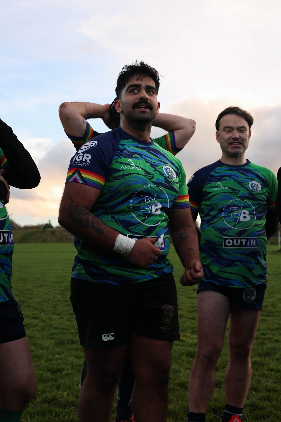 Rugby players in colorful uniforms standing on a field under a partly cloudy sky.