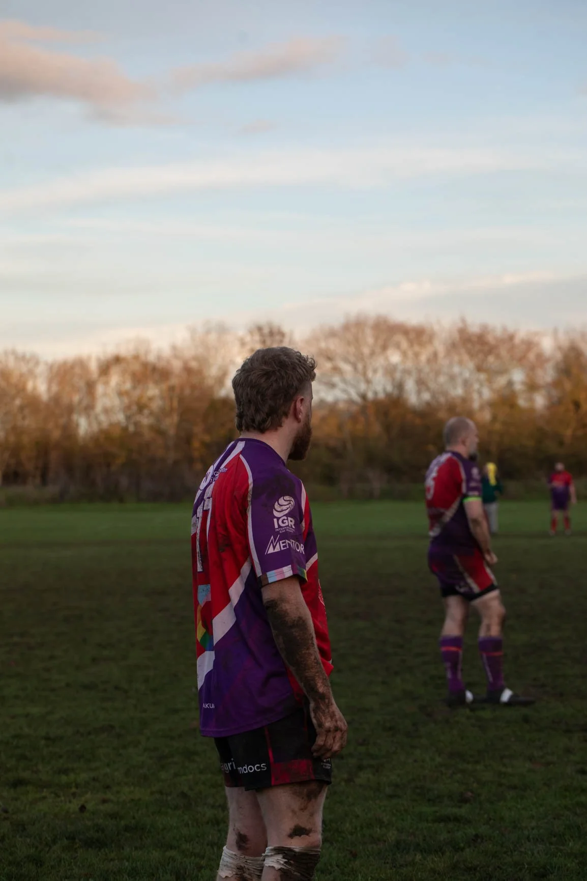 Rugby players on a field during sunset, with one player in the foreground and others in the background, wearing colorful jerseys.