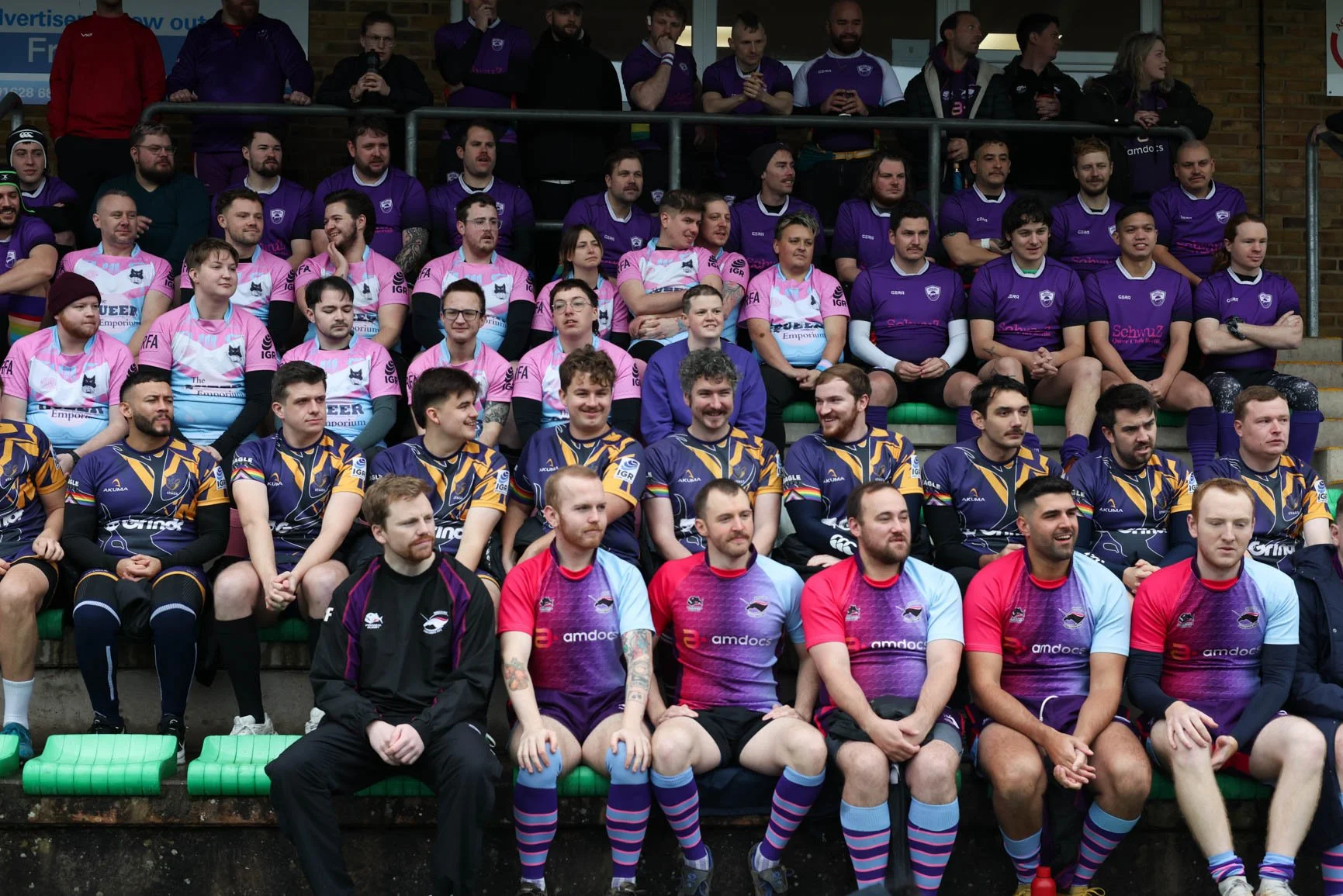 A group of rugby players in colorful jerseys sitting on a stadium bench, with some spectators standing behind them.