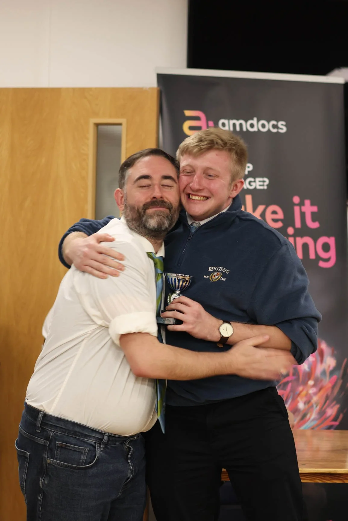 Two men hugging and smiling at an award ceremony, one holding a trophy. The man on the left has a beard, wears a white shirt and a tie, while the man on the right has blonde hair and wears a dark sweatshirt and a wristwatch. Background features a woo