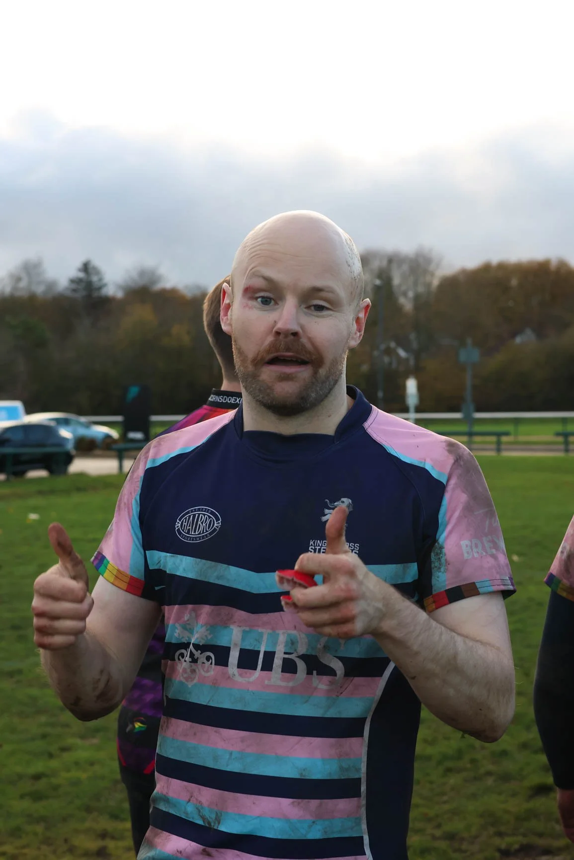 A man with a shaved head and light facial hair wearing a colorful sports jersey, giving a thumbs-up, outdoors on a cloudy day with trees and parked cars in the background.