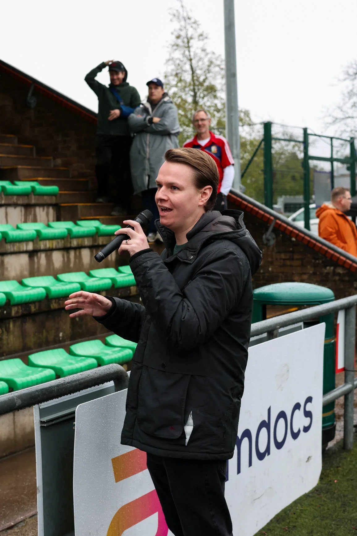 A man in a black jacket speaking into a microphone at a sports event, with a few spectators standing on the steps behind him and a logo sign in the foreground.