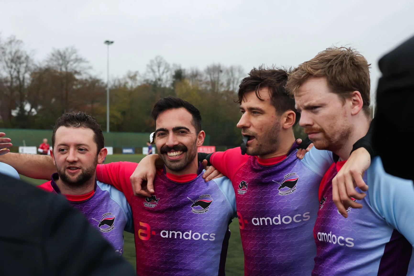 Four rugby players in purple and light blue jerseys huddle together on a field, smiling and celebrating after a game, with trees and a cloudy sky in the background.