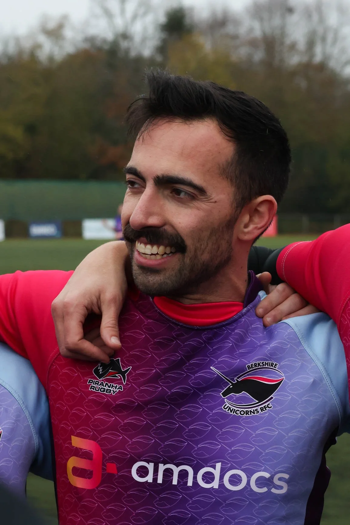 A man with dark hair and a beard smiling, wearing a rugby jersey with logos of Berkshire Unicorns RFC, Piranha Rugby, and AMDOCs, outdoors on a field with trees in the background.