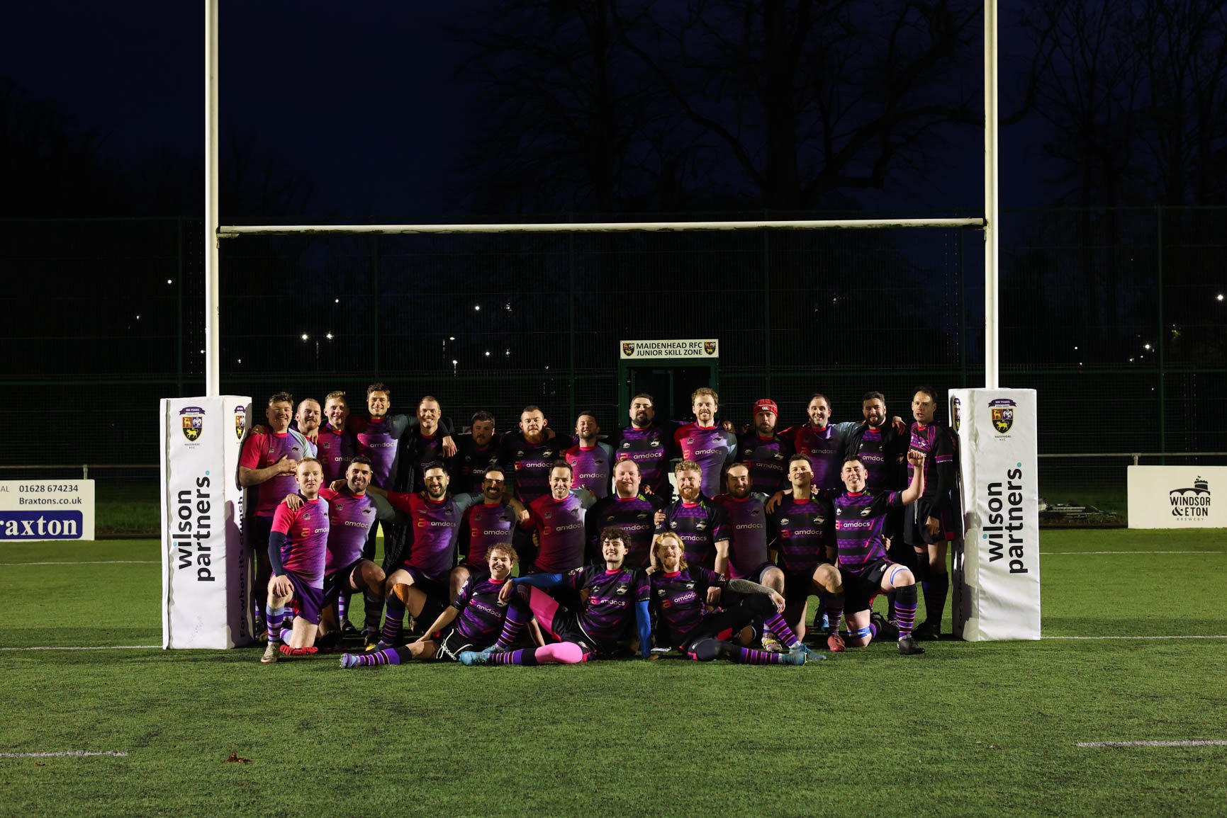 A rugby team wearing purple and black uniforms, posing together on a rugby field at night. They are standing and kneeling in front of rugby goalposts, with some players making gestures or holding a trophy. The background shows darkness and silhouette