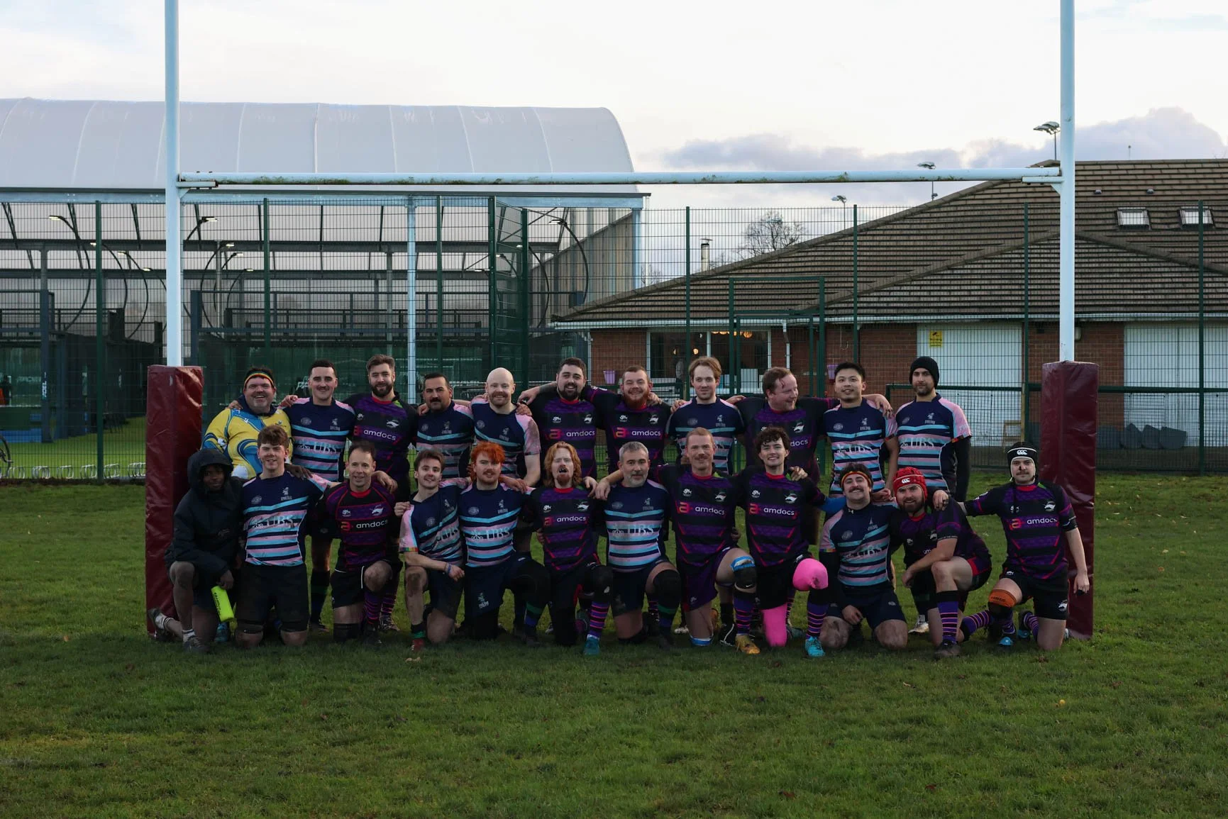 A group of rugby players and coaches posing on a rugby field after a game, standing in front of the goalposts, with a rugby pitch and a building in the background.