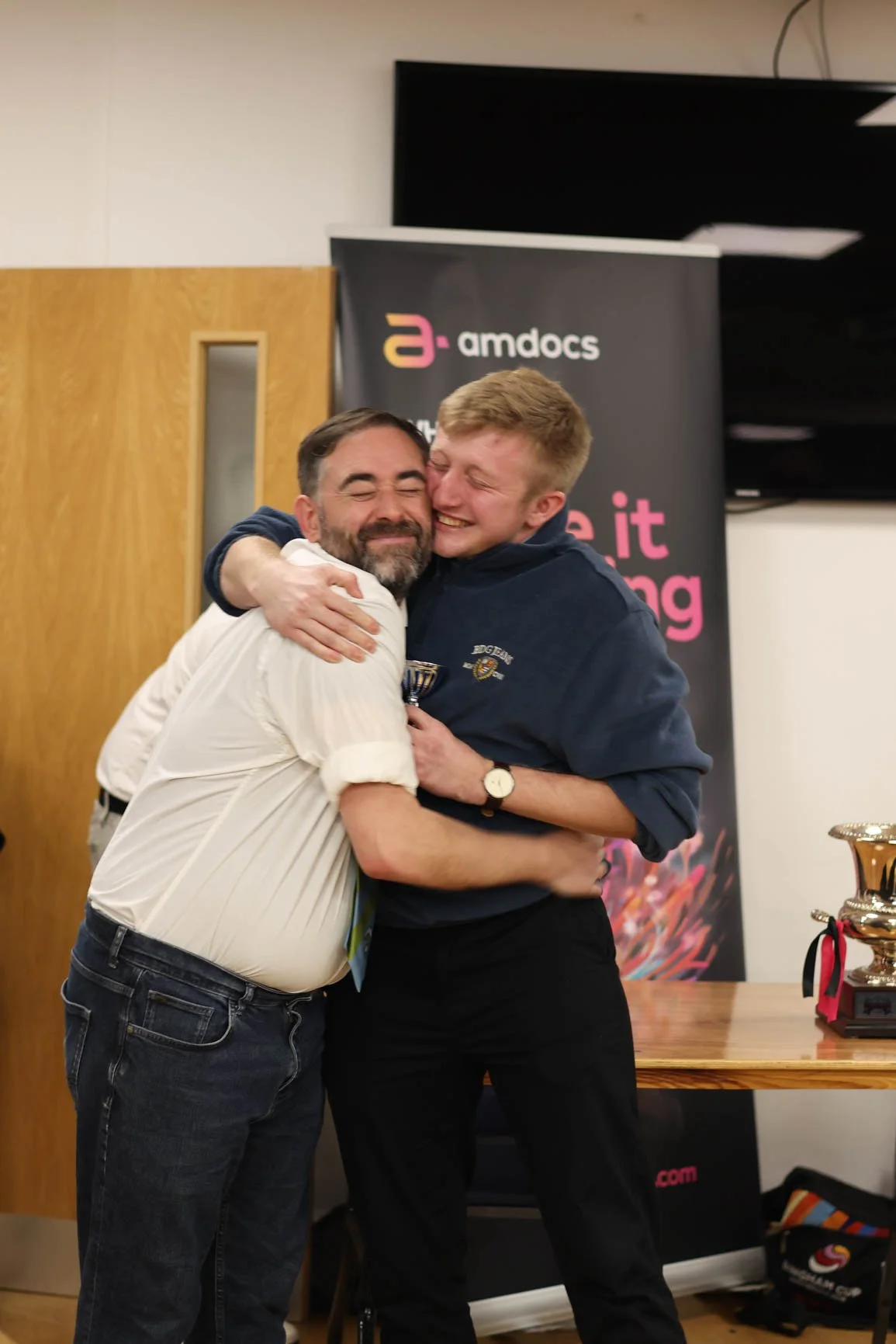 Two men hugging and smiling at an awards ceremony, with a black banner showing 'amdocs' branding in the background.