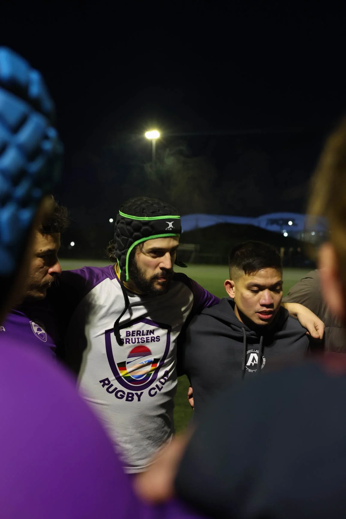 Rugby players huddled together on a field at night, with one player wearing a white jersey with 'Berlin Bruisers Rugby Club' logo and a black headgear, during a team meeting or pep talk.