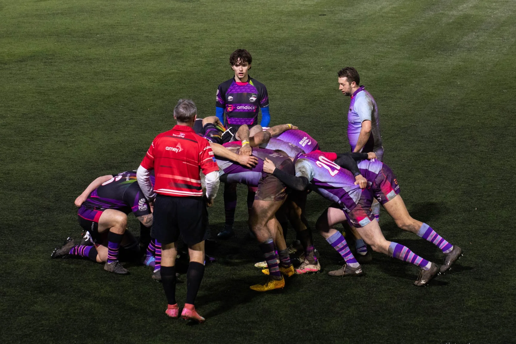 Rugby players engaged in a group scrum on a grassy field, with referees observing.