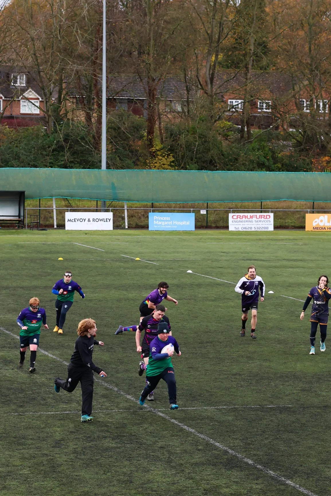 Youth rugby players running on a field during practice or game, with autumn trees and advertising banners in the background.