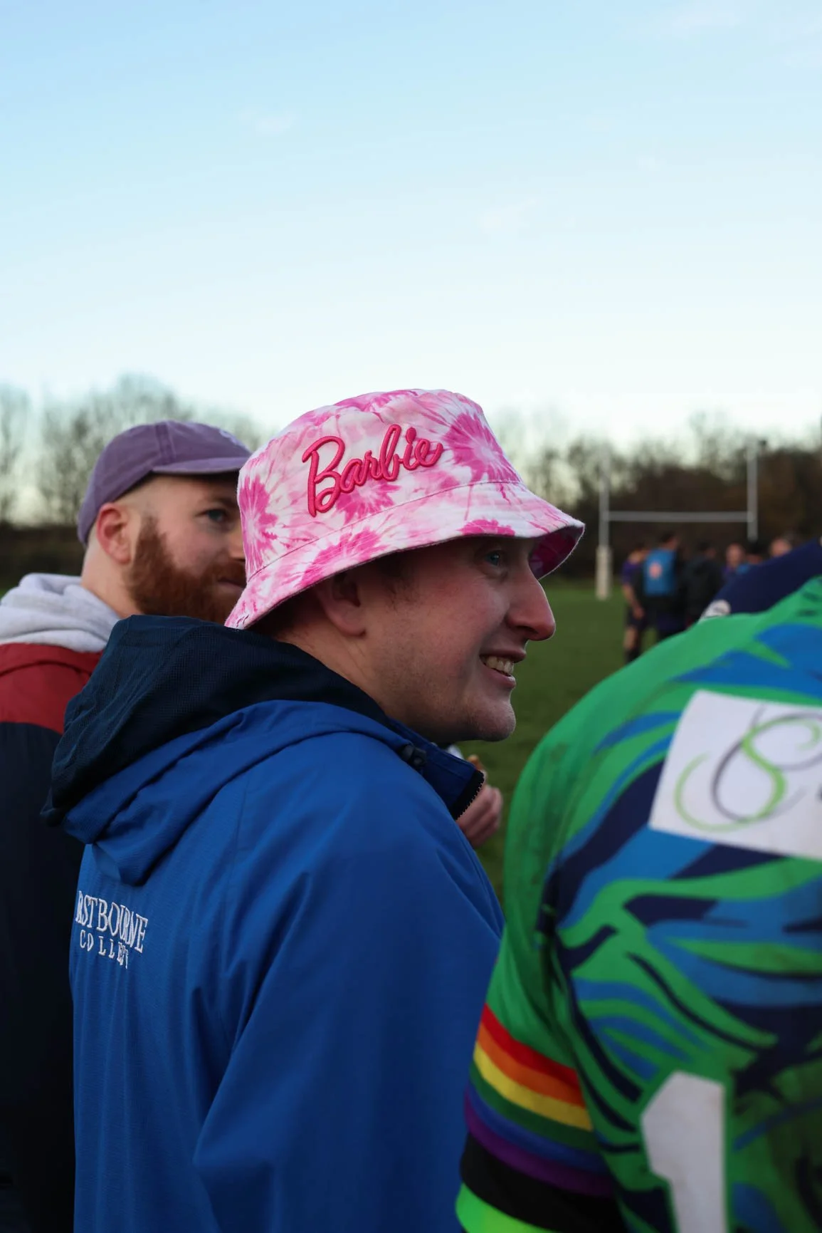 A group of people outdoors, with a man wearing a pink tie-dye bucket hat with the word 'Barbie' in pink embroidery, smiling and talking to others.
