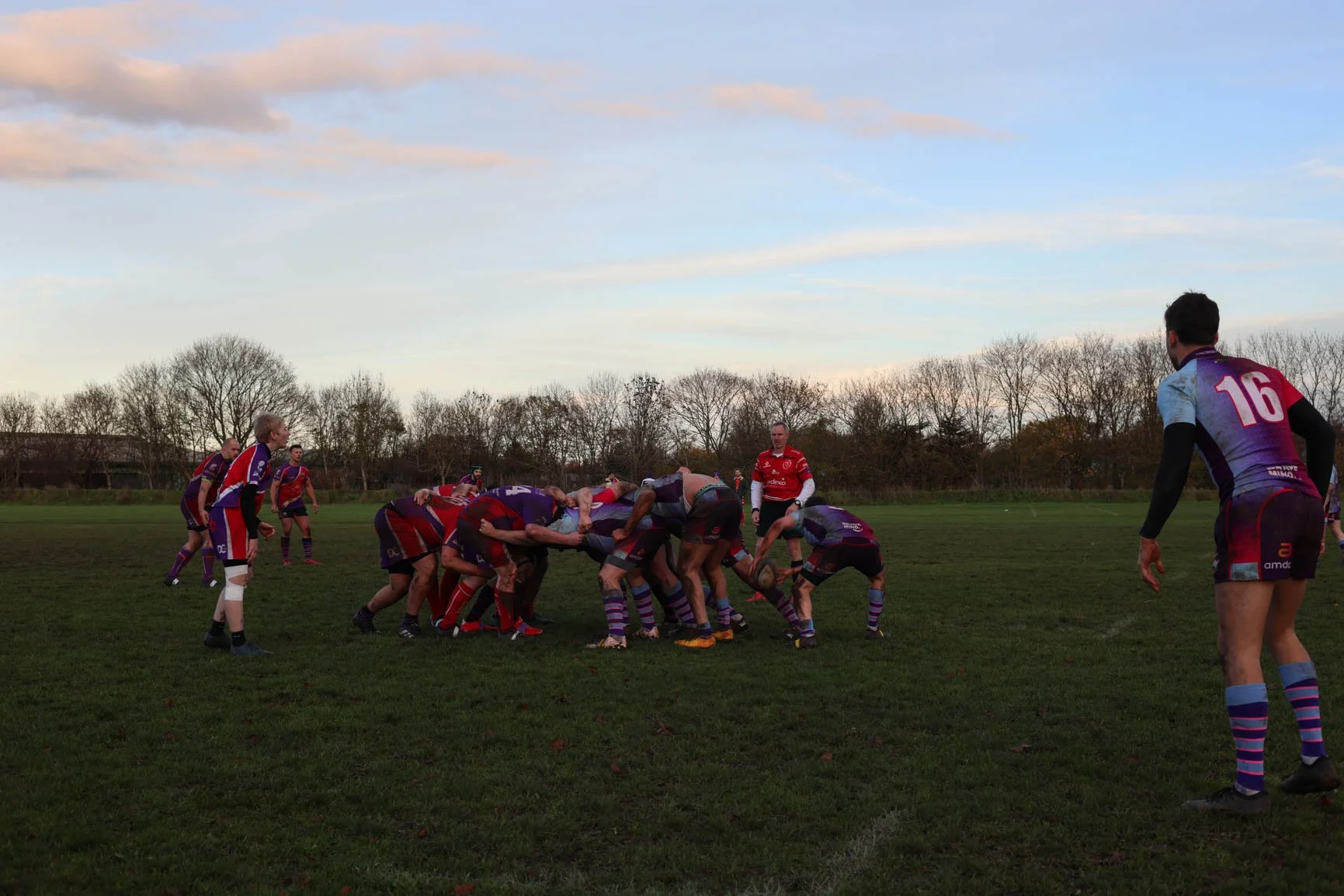 Rugby game with players engaged in a scrum on a grassy field during late afternoon; some players are bent over with hands on the ground, wearing colorful jerseys and shorts, with a few players standing nearby, and trees and a partly cloudy sky in the